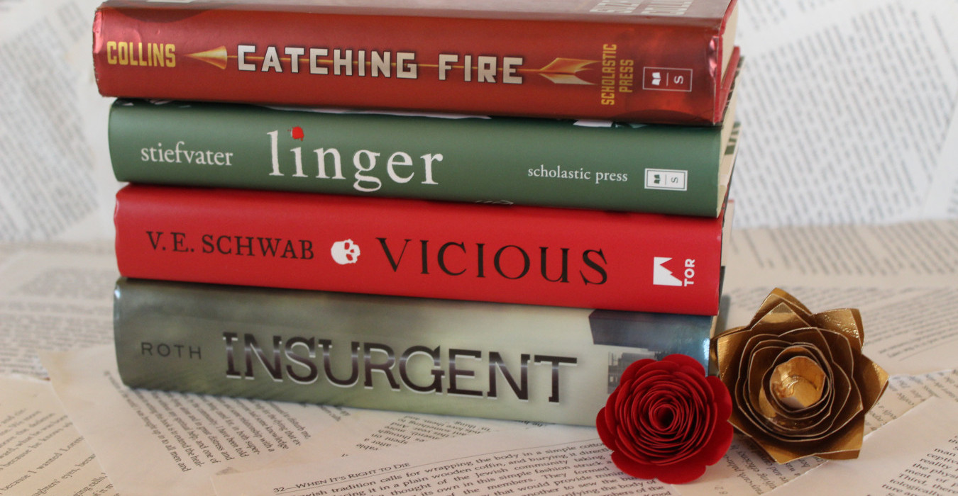 A stack of red and green books. Red and gold paper flowers sit at the bottom of the stack. From top to bottom, the books are Catching Fire, Linger, Vicious, and Insurgent