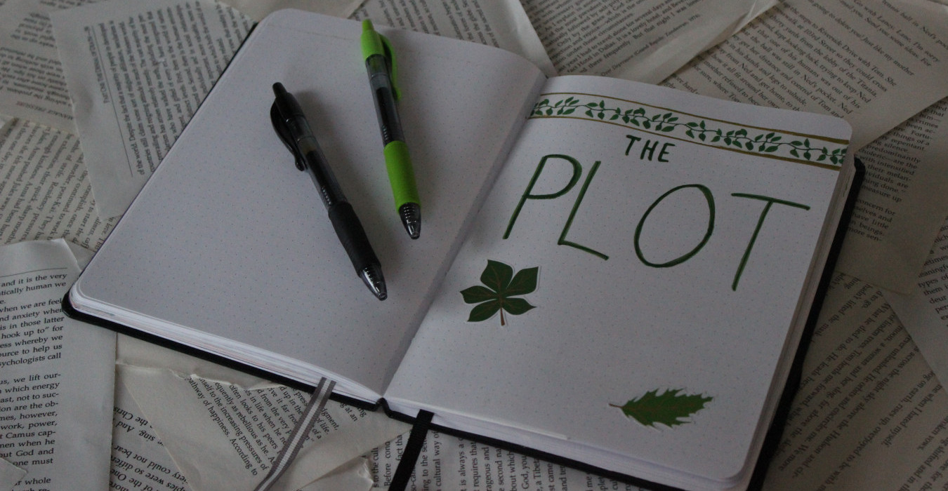 An open notebook that reads "THE PLOT" in large bold letters. The page is decorated with stickers of leaves. Two pens are set on top of it.