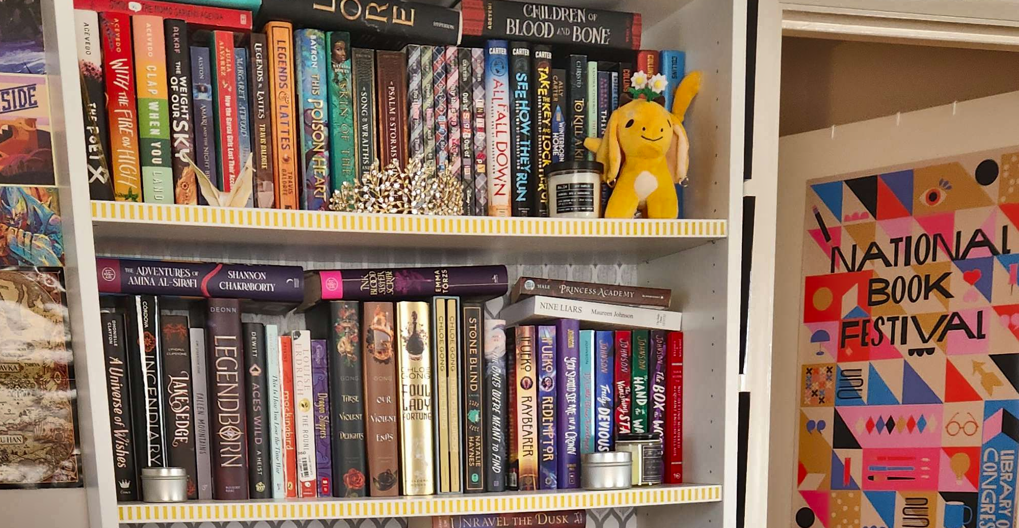 Photograph of two shelves on a tall bookshelf. The books are stacked a bit haphazardly, and there are too many to describe. The shelves are also decorated with a paper crane, a crown, candles, and a plush toy. In the background is an open door, which is decorated with a poster for the National Book Festival in Washington DC.