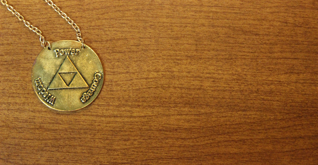 A wooden background with a gold pendant necklace on top. The pendant shows the triforce from The Legend of Zelda. Around it are the words Power, Courage, Wisdom.