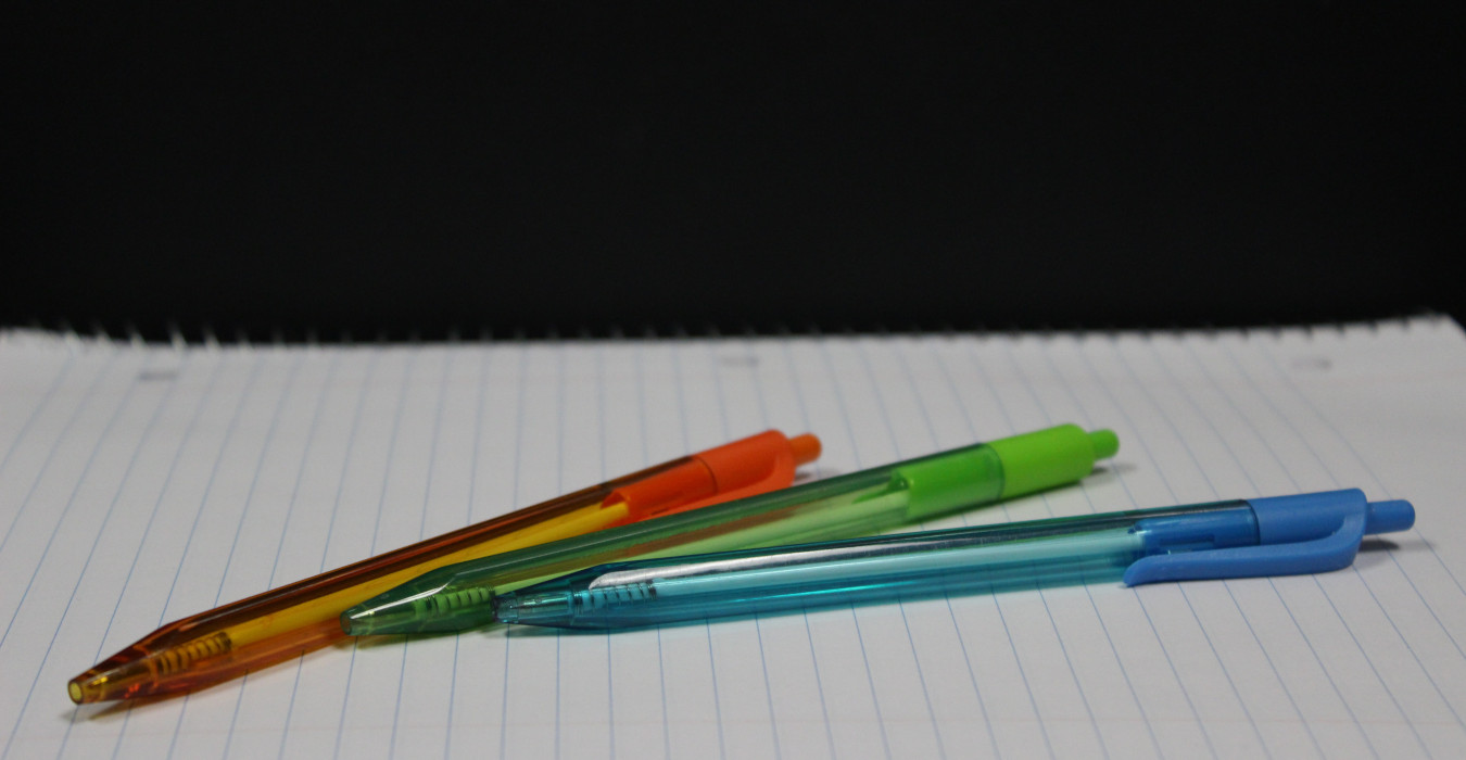 An orange, green, and blue pen sit on a blank notebook.