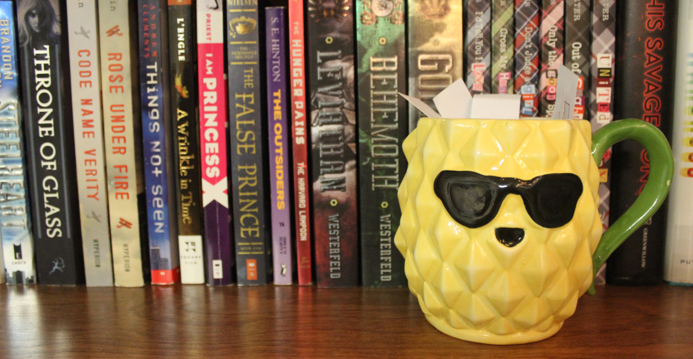 A large mug shaped like a pineapple sits on a bookshelf with several slips of paper sticking out of it. The pineapple is wearing sunglasses