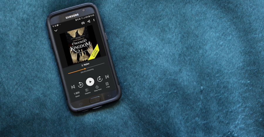 A cell phone displaying the Crooked Kingdom audiobook. The phone sits against a dark teal background.