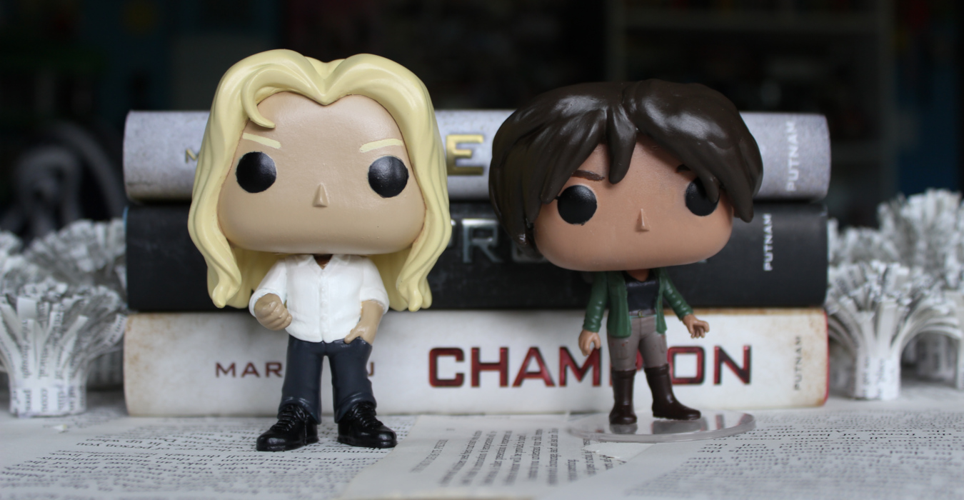 Two funko pop figurines depicting Day and June from Legend by Marie Lu. The figurines are standing in front of a stack of the Legend trilogy.