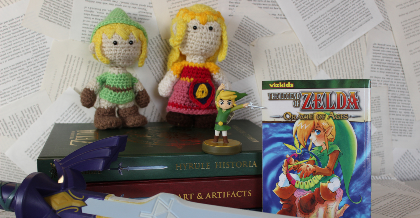 A display of Legend of Zelda books and various memorabilia. In the back is a stack of books from the "Goddess Collection," which includes Hyrule Historia, Art & Artifacts, and the Encyclopedia. Stacked on top are two crocheted dolls of Link and Zelda, along with an amiibo figurine based on Link from The Wind Waker. Beside it is a cop of the Oracle of Ages manga, and in front is a prop replica of the Master Sword.