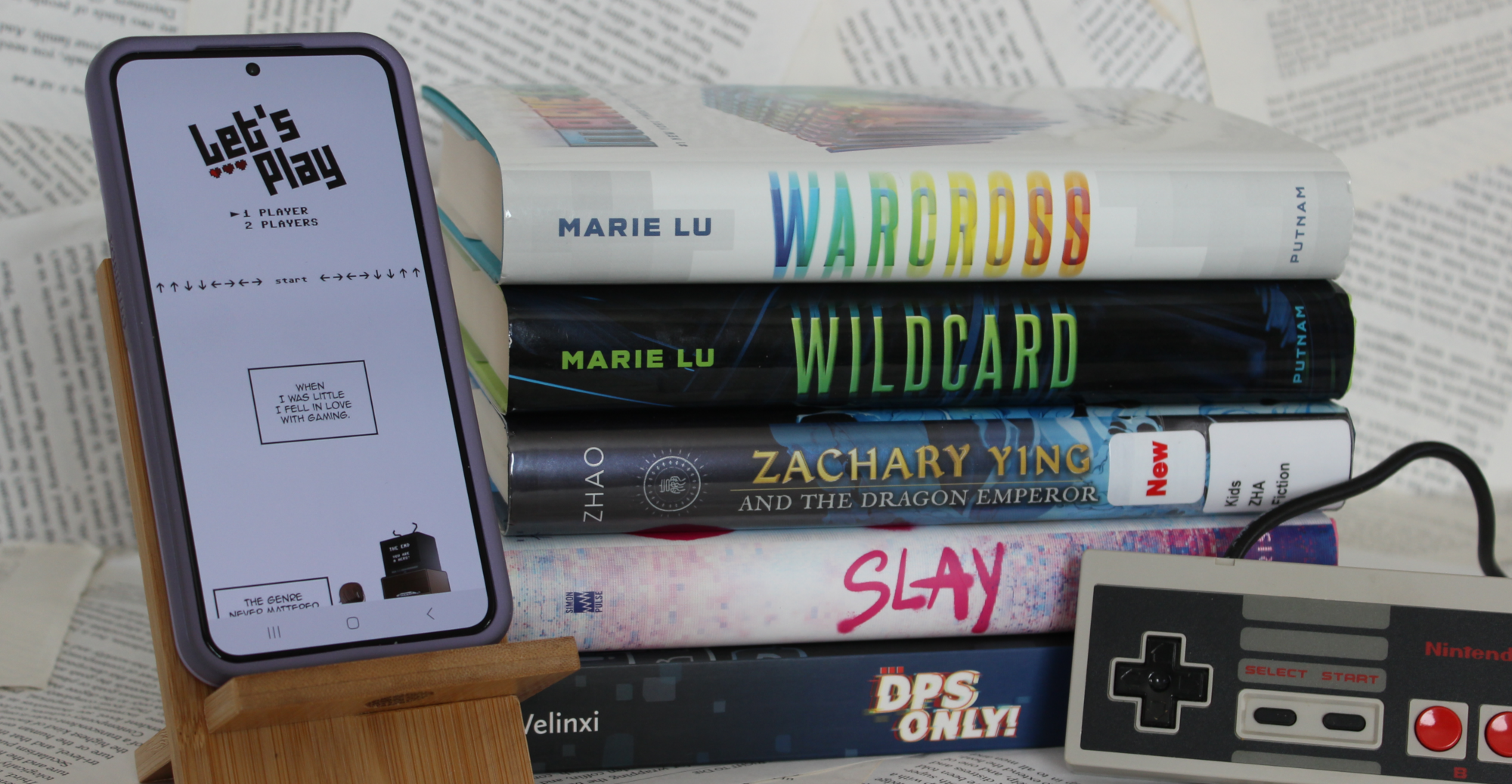 Photo of a stack of books as well as a smartphone on a wooden stand. The smartphone screen shows the beginning of the webcomic Let's Play. The stack of books in order from top to bottom is: Warcross, Wildcard, Zachary Ying and the Dragon Emperor, Slay, and DPS Only!. Leaned against the stack of books is a rectangular controller for the Nintendo NES Console.