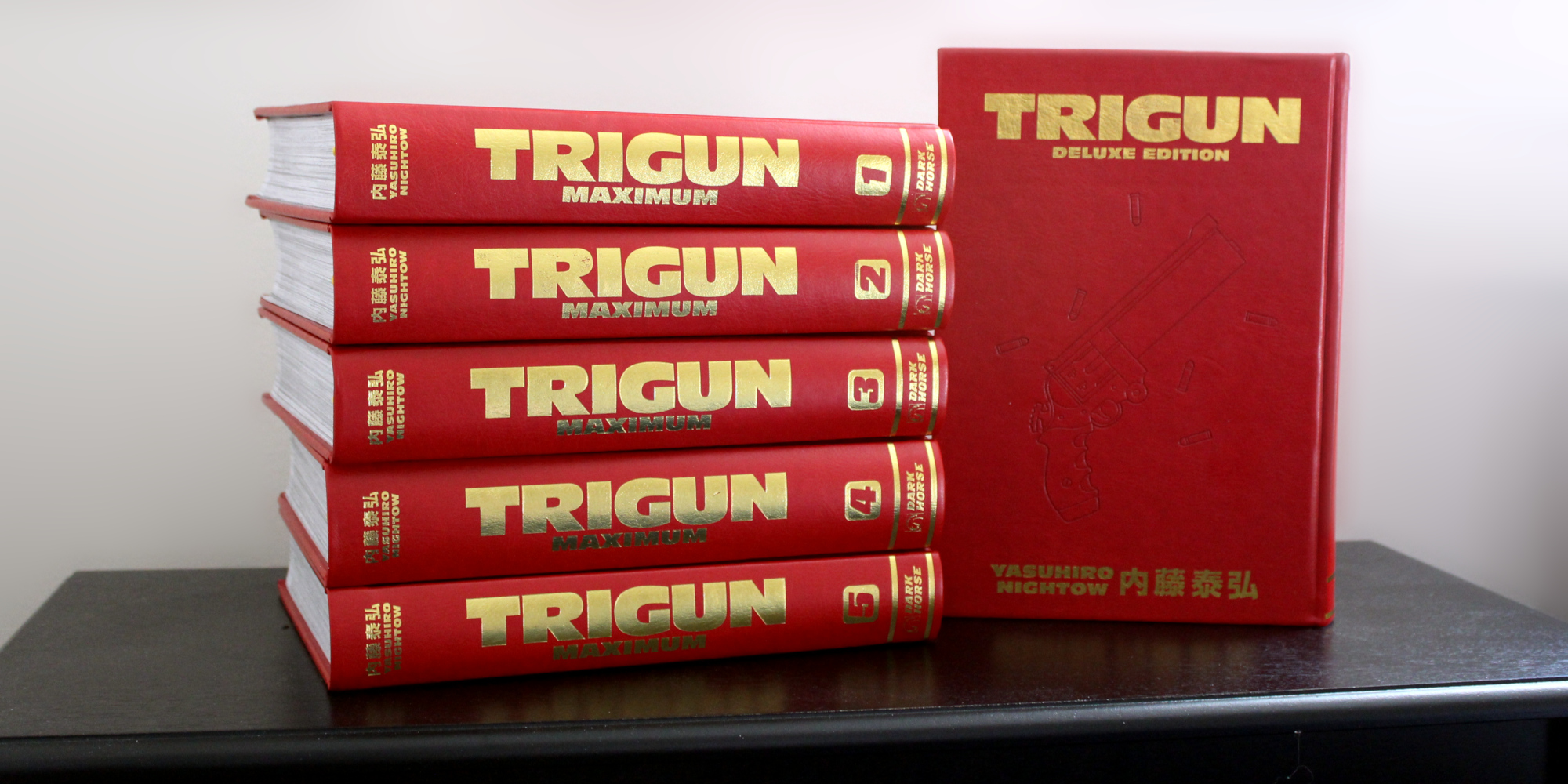 Photo of a collection of the Trigun and Trigun Maximum Deluxe Editions, published by Dark Horse. It's a stack of five thick books with red covers on the left, and another book with a red cover propped up on the right. The cover is embossed with Vash's handgun.